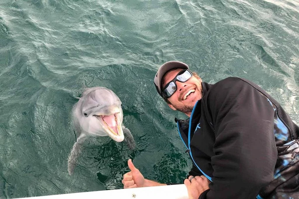 Man leaning over the edge of the boat to be near a dolphin the water. Both of them are smiling and looking at the camera