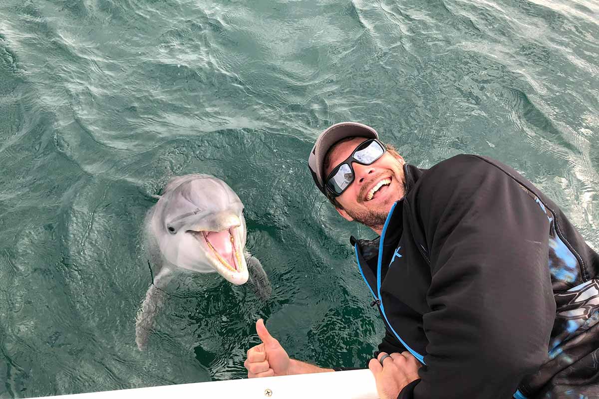 Man leaning over the edge of the boat to be near a dolphin the water. Both of them are smiling and looking at the camera