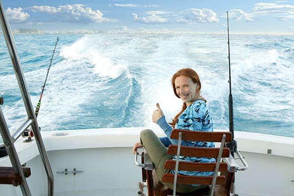 Woman fishing from back of a charter boat. She's turning her head around holding a thumbs up and is smiling