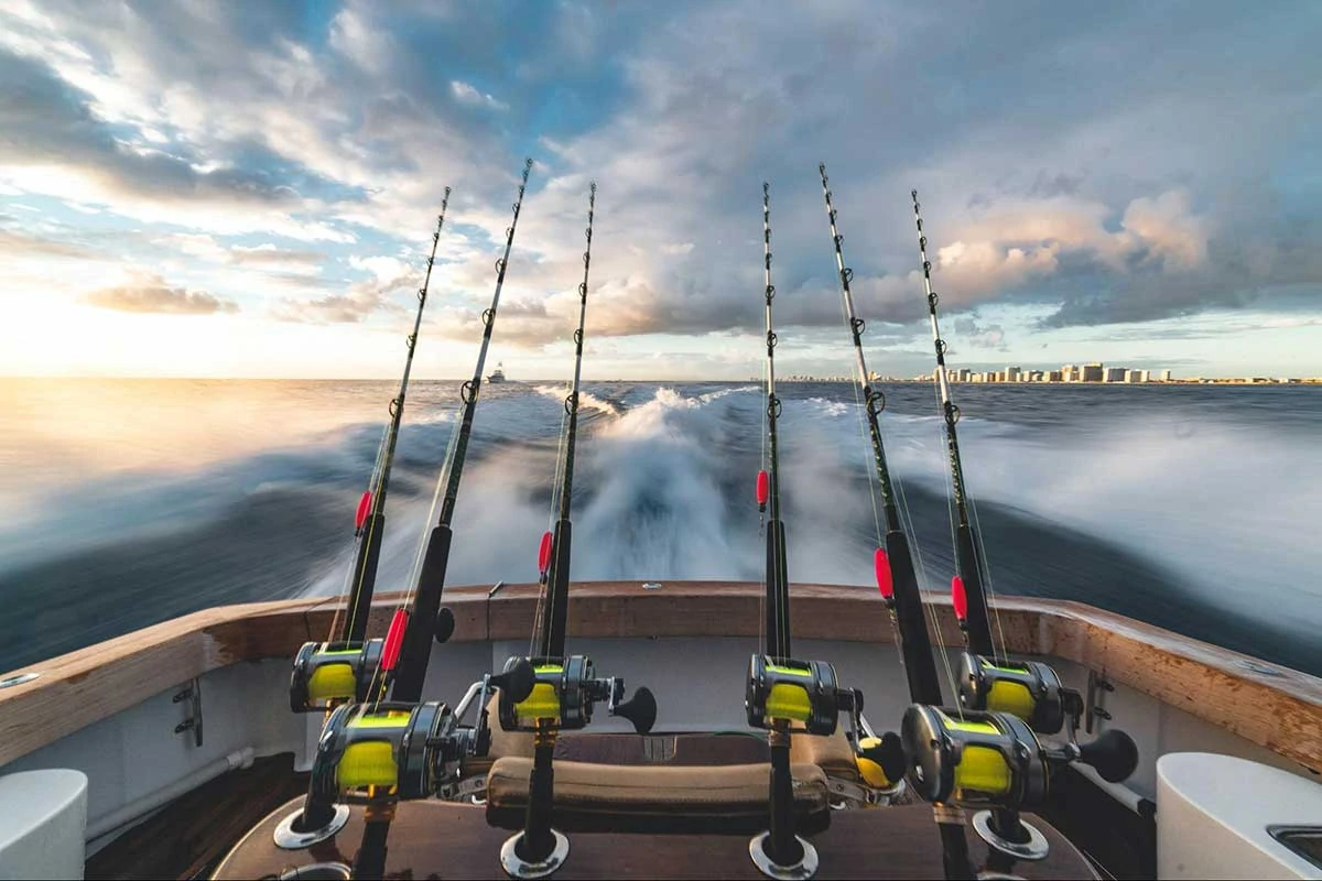 Photo of fishing boat taken from the bow, facing the stern. The back of the boat is visible with 6 fishing poles and the wake of the boat is blurry from the speed. The sky is partly cloudy and looks expansive and beautiful.