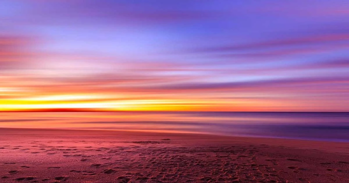 Photo that looks like a painting of the sky at sunset. The beach is in the foreground with footprints in the sand, and the water, sunrise, and dawning sky look like paint brushings across the page.
