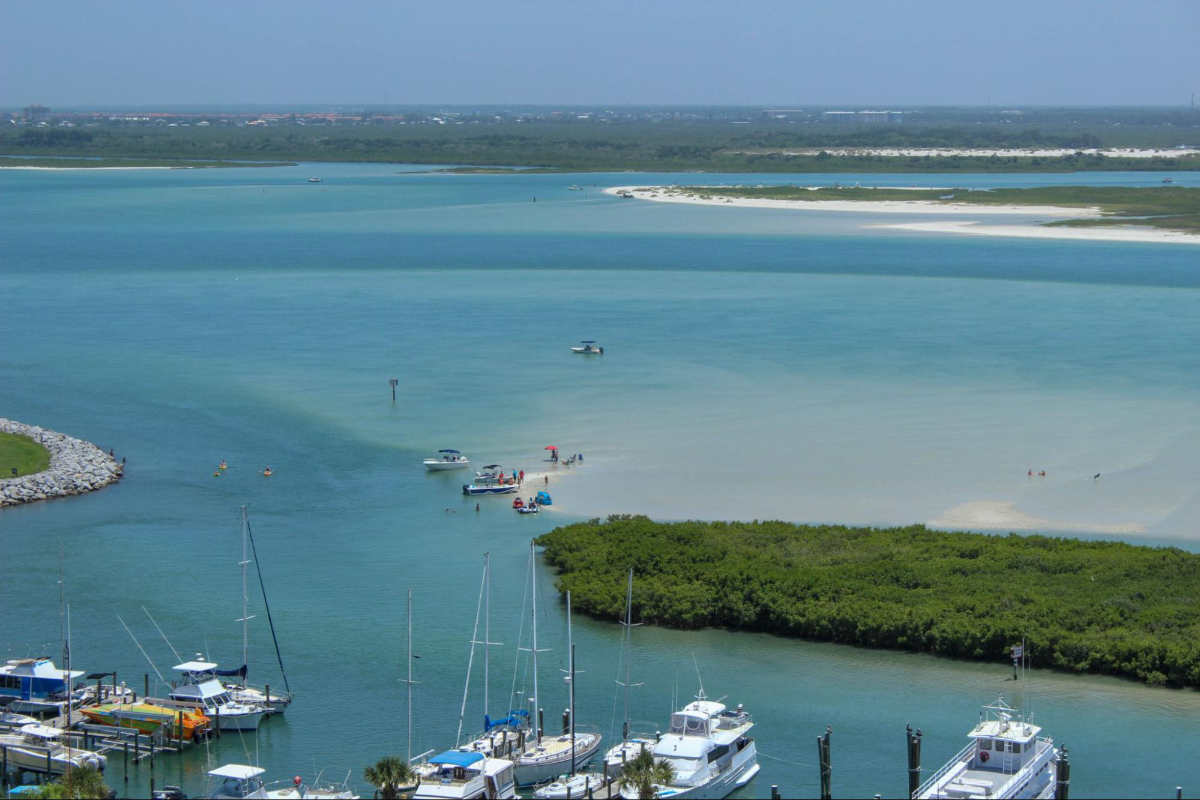 Aerial photo of boats at a marina at an inlet. There is a sandbar by one of the barrier reefs where people are standing in the water.