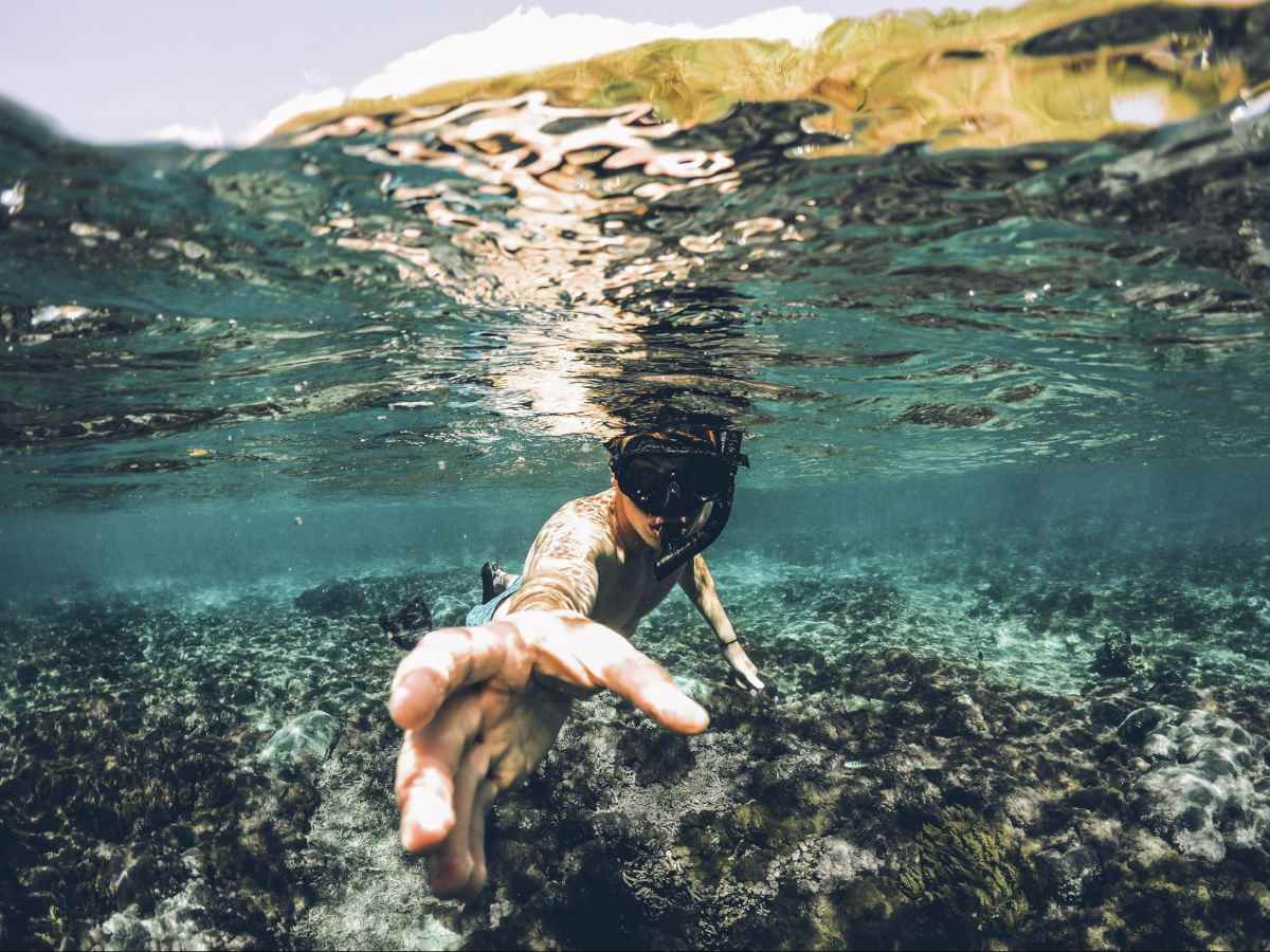 Photo of young man snorkeling. The photo is shot from half in the water and half out. The boy's hand is extended as if to grab something towards the camera.